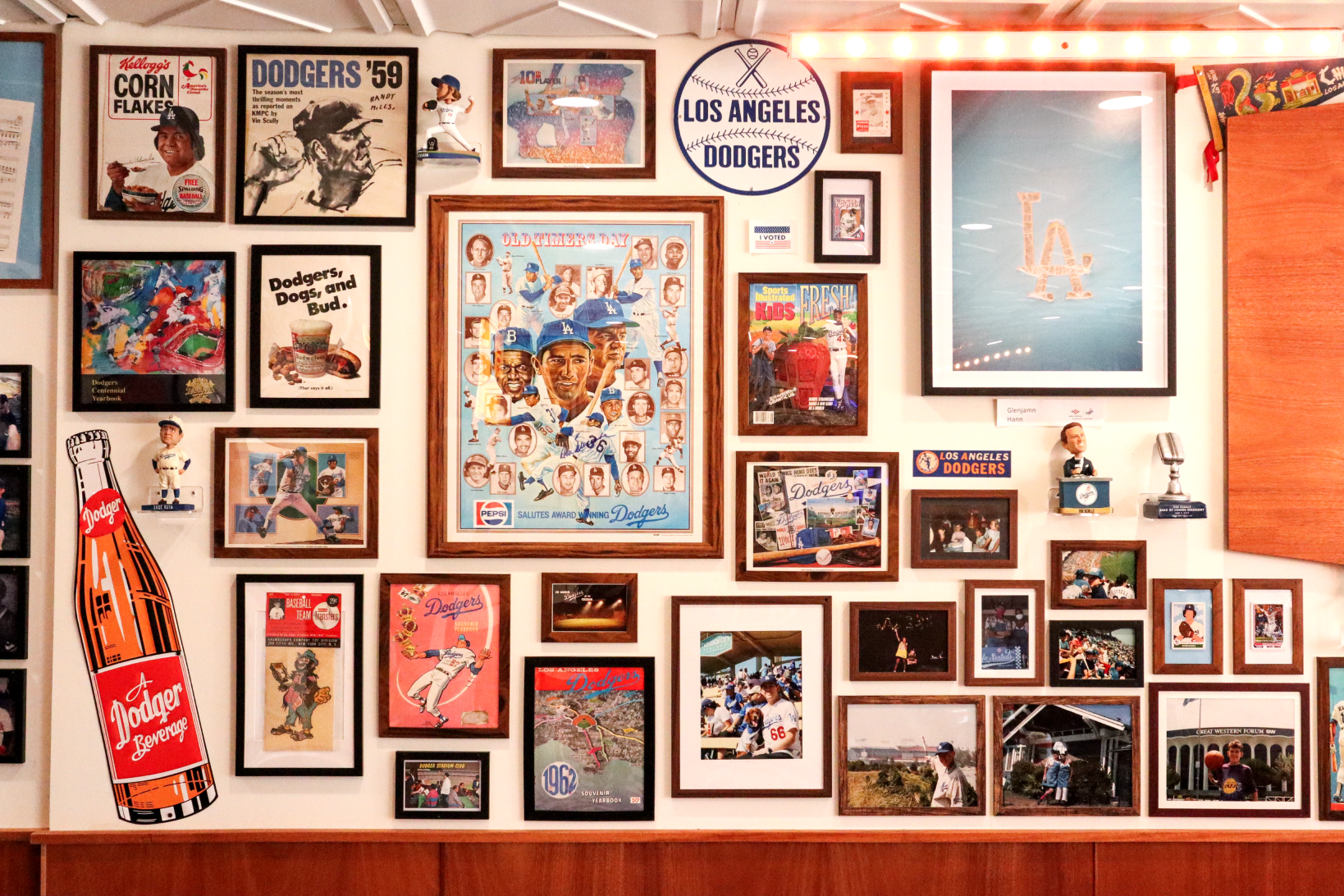 Wall filled with framed Los Angeles Dodgers memorabilia including posters, photos, bobbleheads, and a vintage Dodgers beverage bottle cutout.